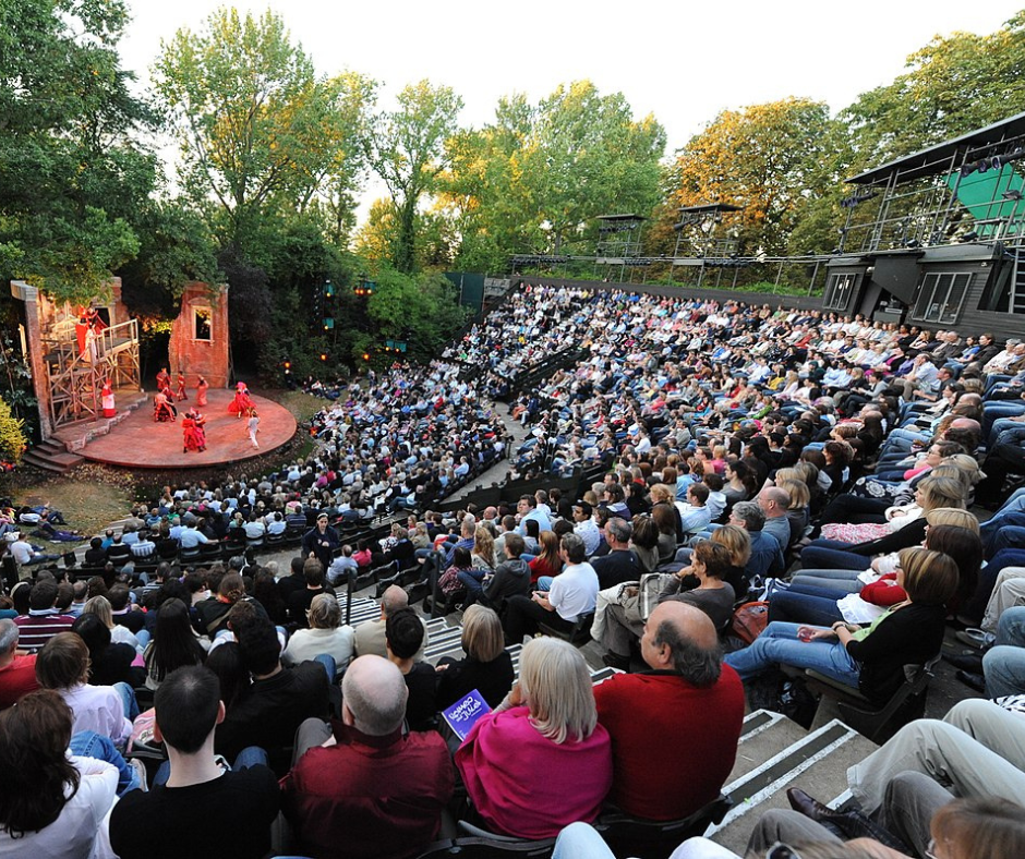 Tiered seating full of people watching performers on an open air stage in the park