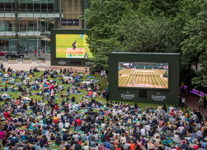 Wide shot of people sitting on the grass, in front of a big screen in the park.